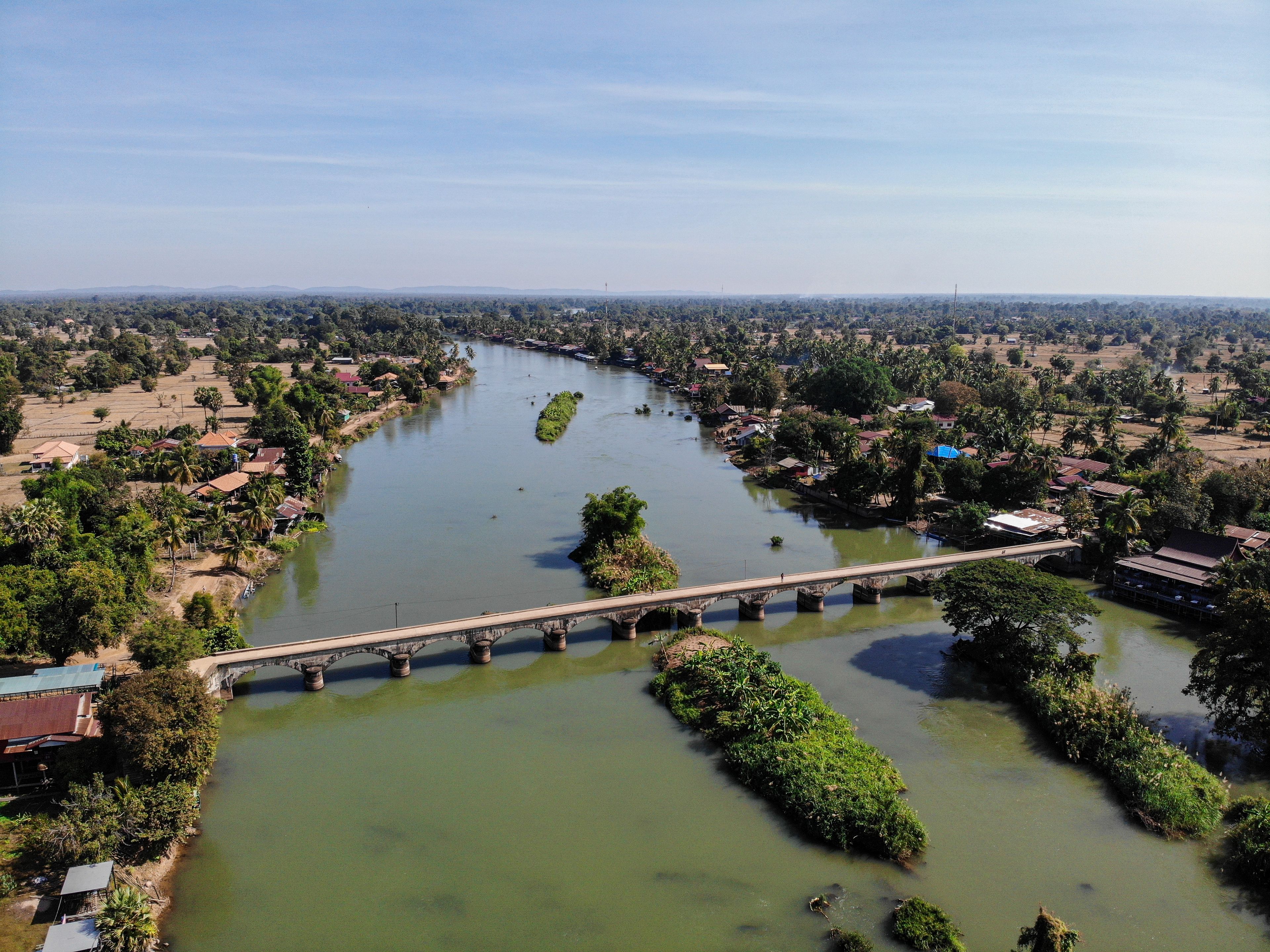 Um rio com uma ponte, rodeado por vegetação e edifícios baixos.