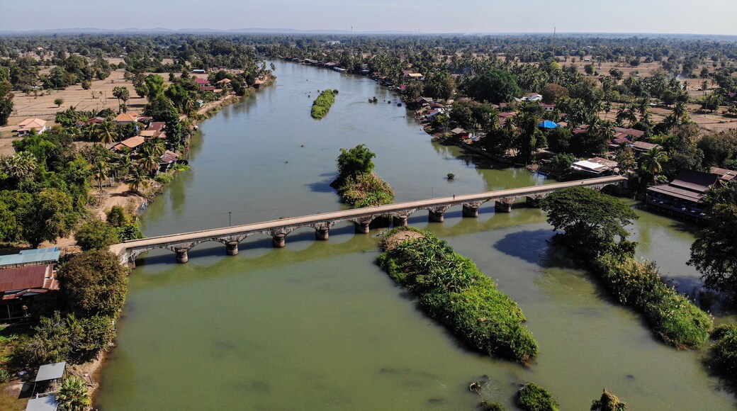 Um rio com uma ponte, rodeado por vegetação e edifícios baixos.
