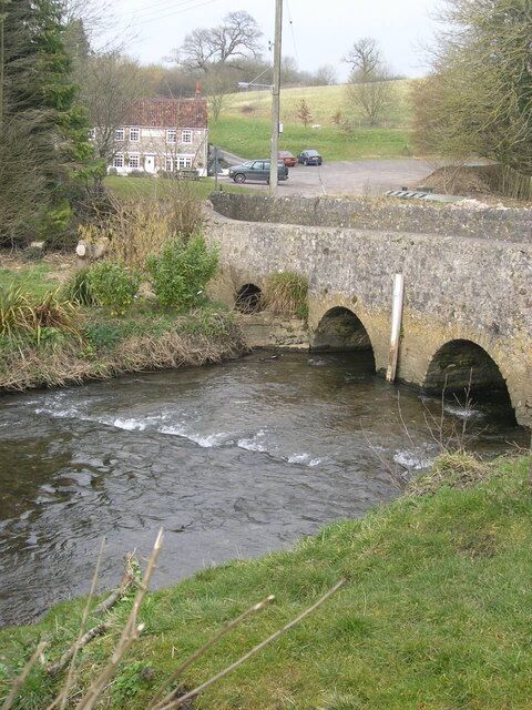 A stone bridge over a flowing stream with a house and cars in the background.