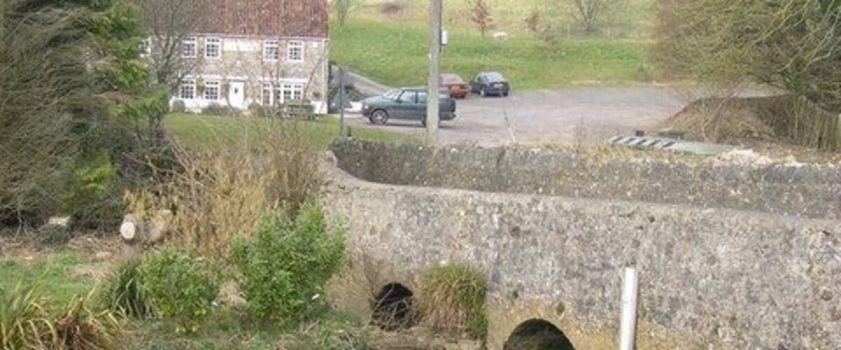 A stone bridge over a flowing stream with a house and cars in the background.