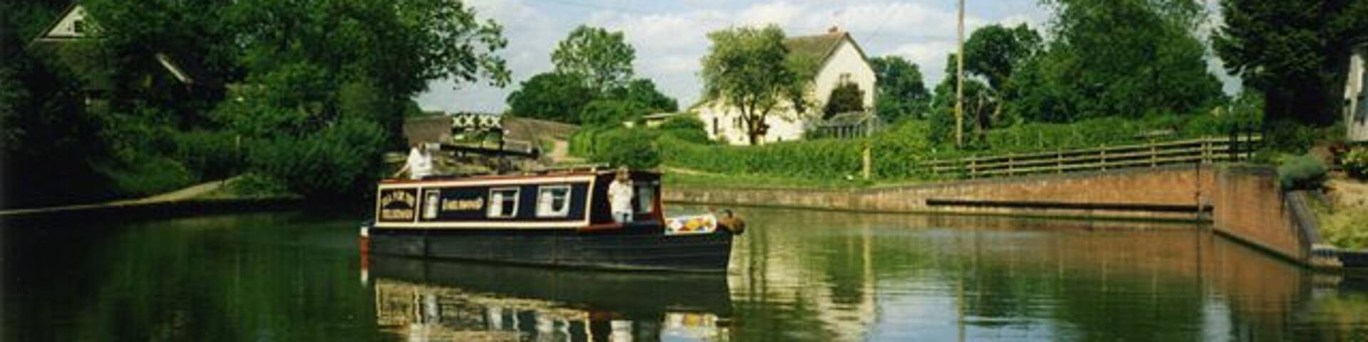 A narrowboat on a calm river with a bridge and greenery in the background.
