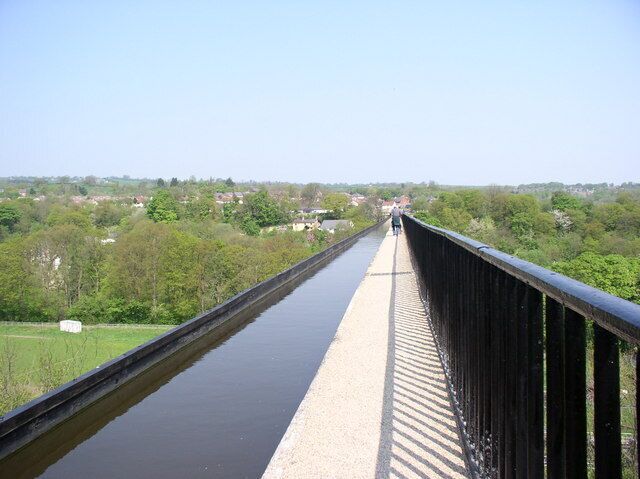 A long bridge with a water channel on one side, surrounded by greenery and distant buildings.
