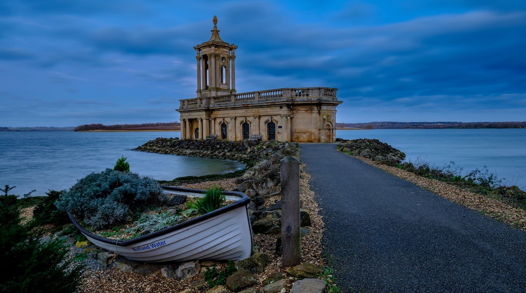 A historic building with a tower by a lake, a boat labeled 'Rutland Water' on the shore, and a pathway leading to the structure.