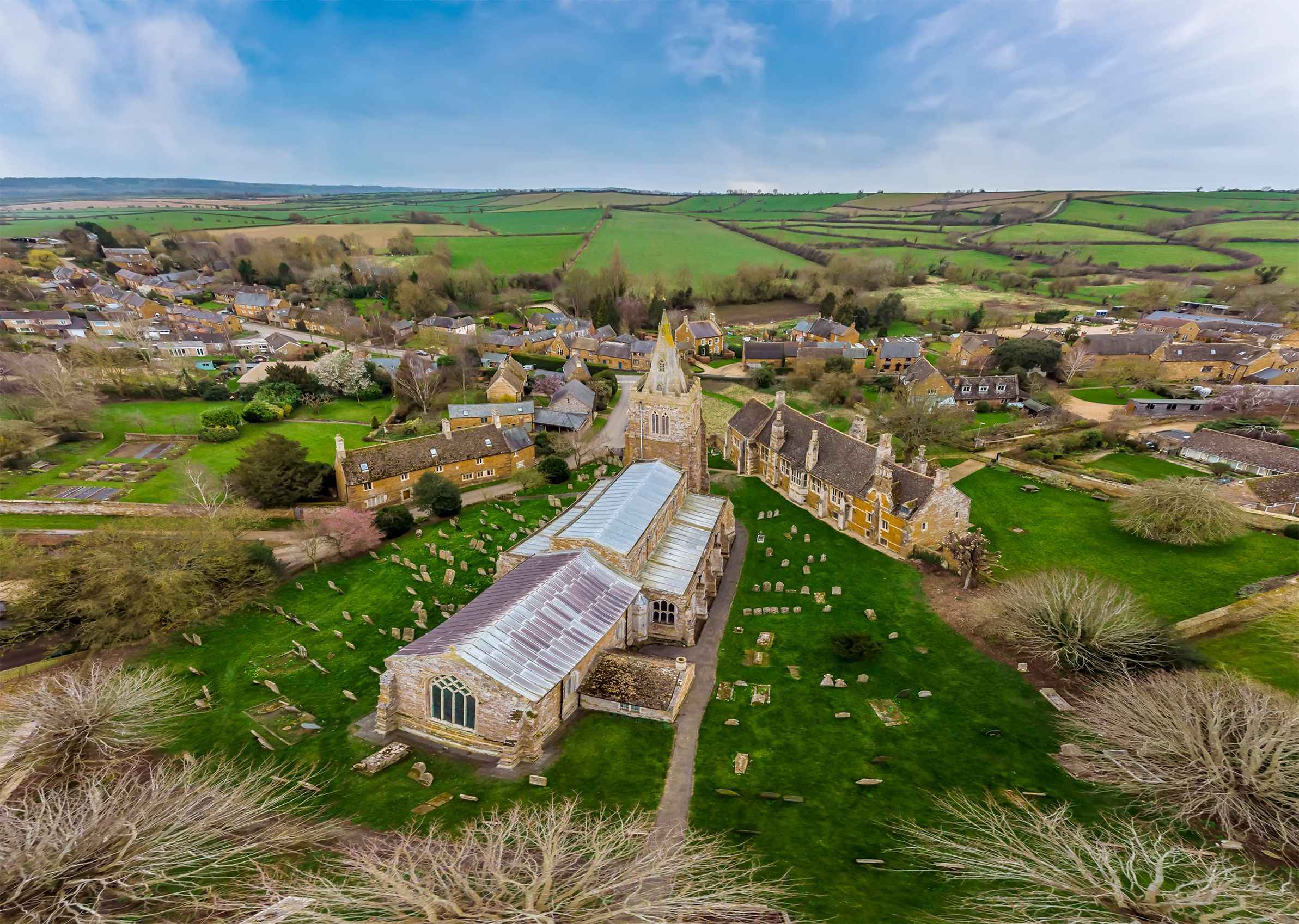 A village with a prominent church featuring a large stained glass window, surrounded by green fields and scattered houses.