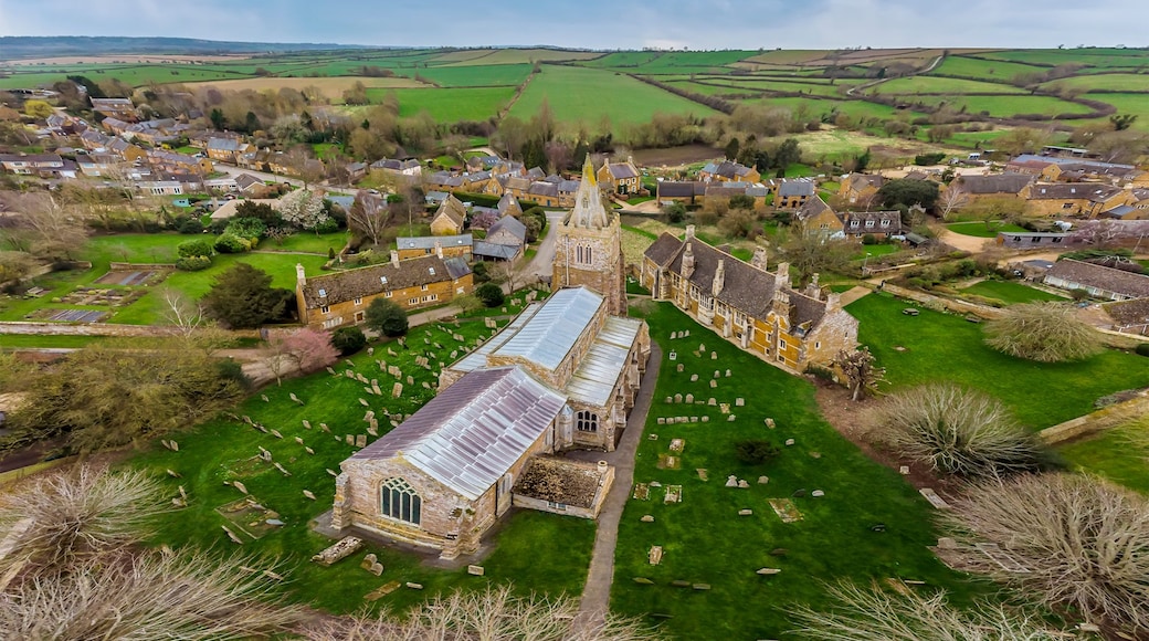 A village with a prominent church featuring a large stained glass window, surrounded by green fields and scattered houses.