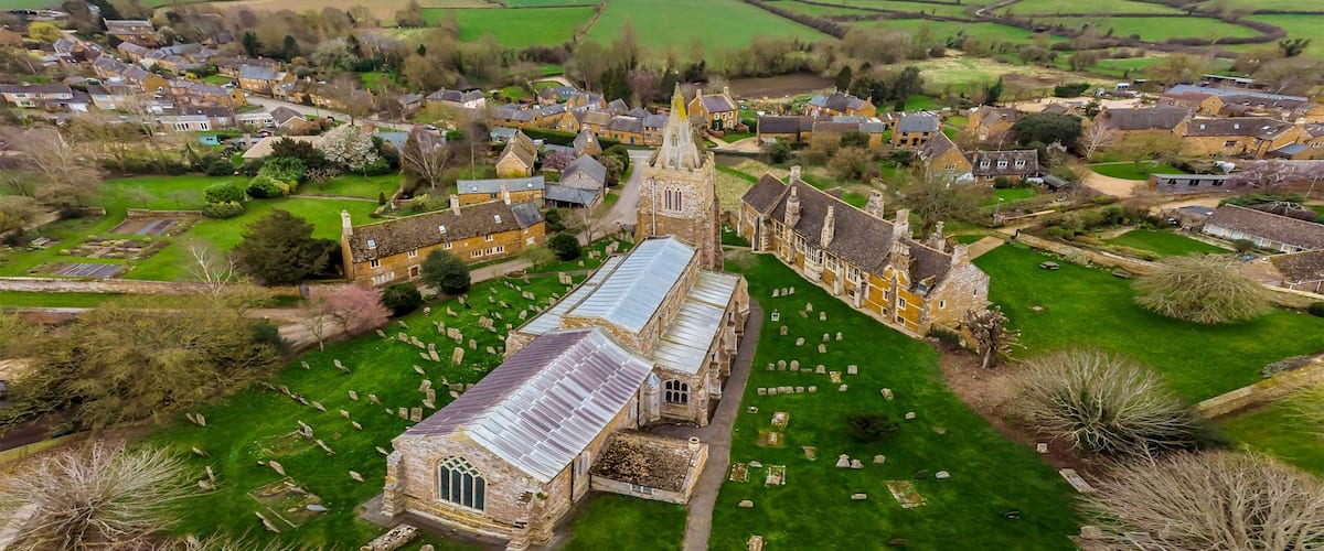 A village with a prominent church featuring a large stained glass window, surrounded by green fields and scattered houses.