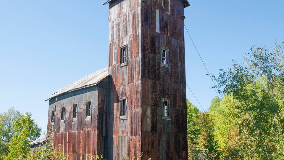 A large, rusted, wooden building with multiple windows and a tall tower, surrounded by trees and a chain-link fence.