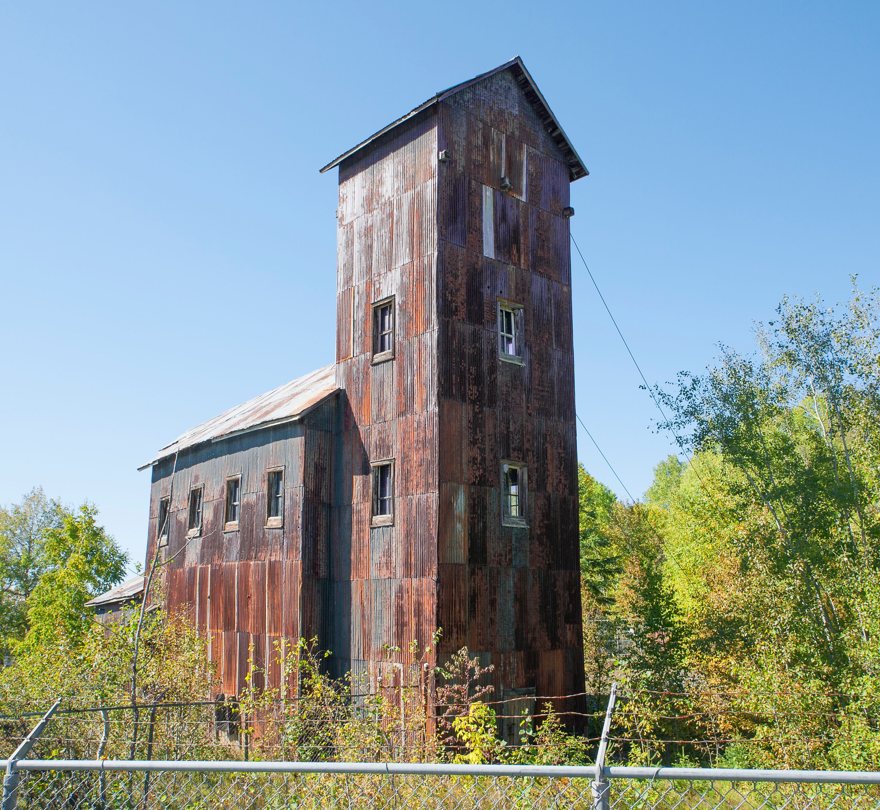 A large, rusted, wooden building with multiple windows and a tall tower, surrounded by trees and a chain-link fence.