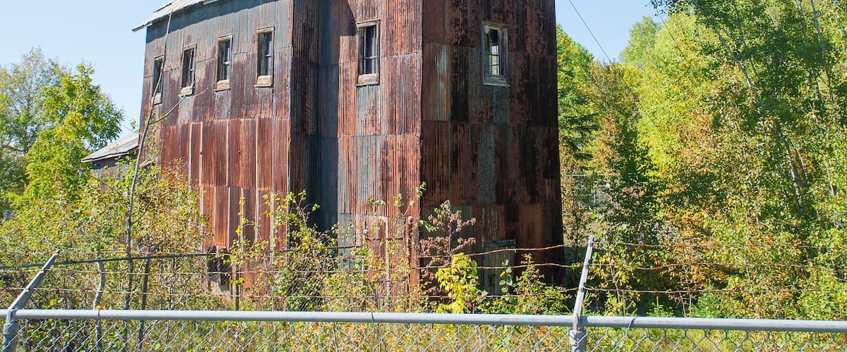 A large, rusted, wooden building with multiple windows and a tall tower, surrounded by trees and a chain-link fence.
