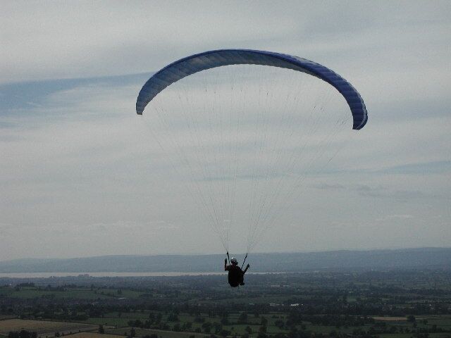 A paraglider in mid-flight over a rural landscape with fields and distant hills.