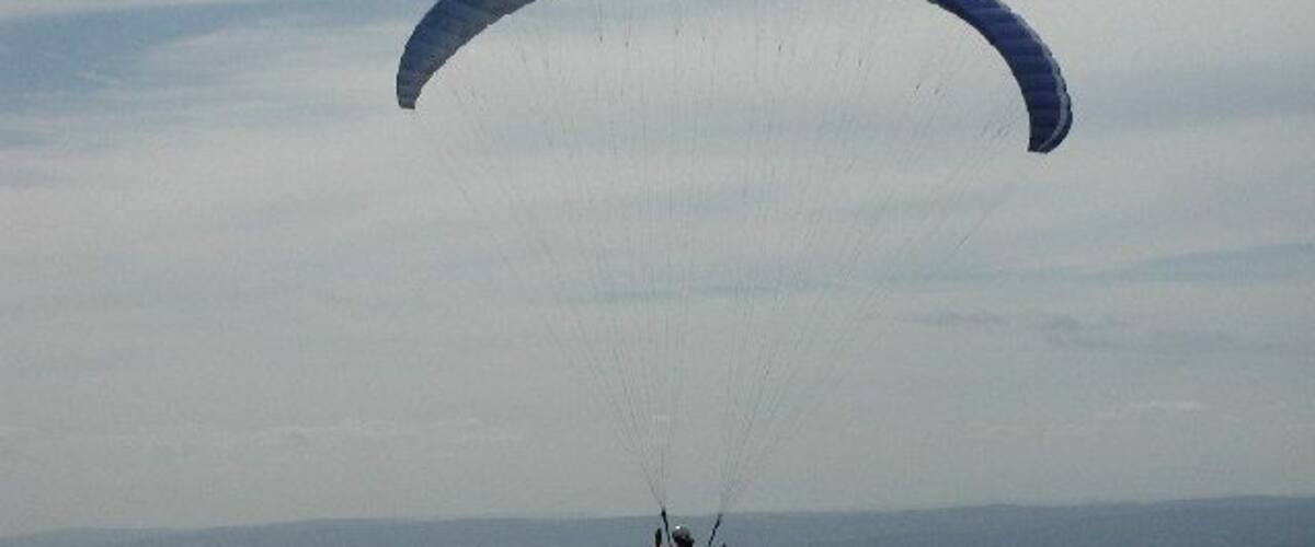 A paraglider in mid-flight over a rural landscape with fields and distant hills.