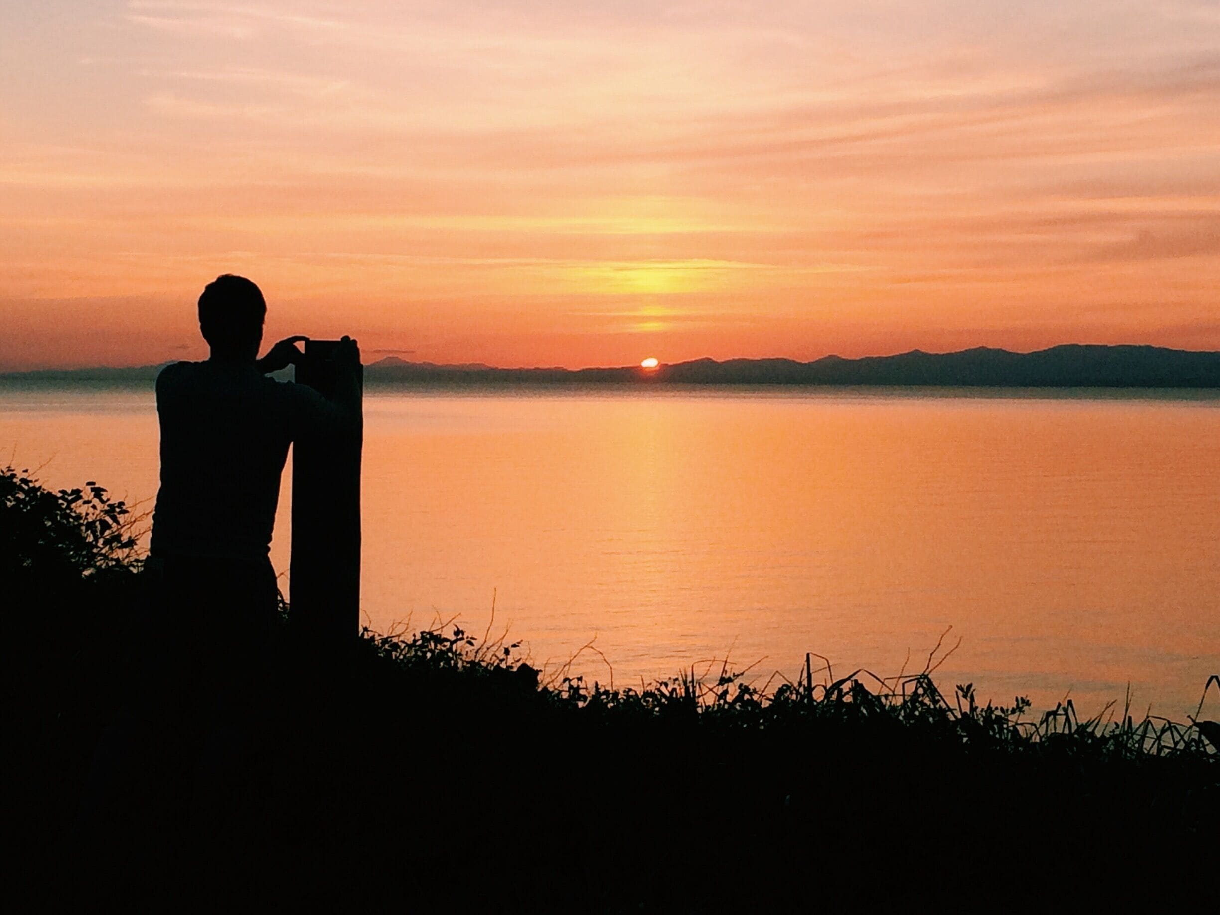 A silhouette of a person sitting by a calm lake at sunset.
