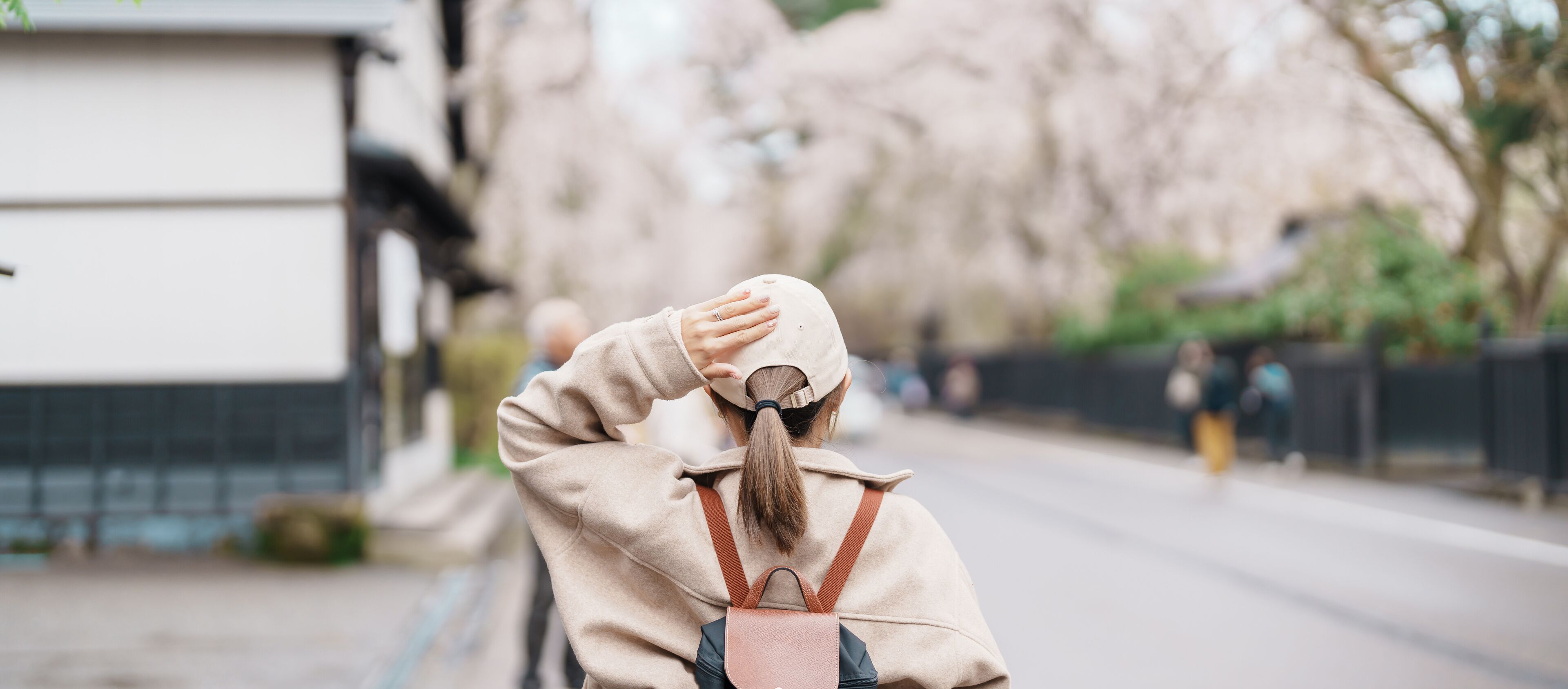 A person in a beige coat and hat walking down a street lined with trees and buildings.