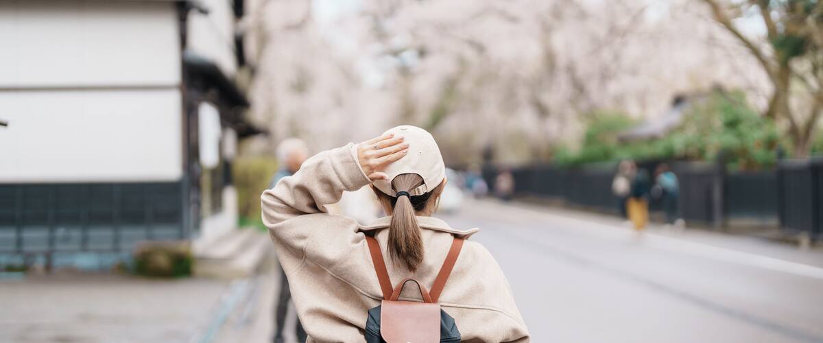 A person in a beige coat and hat walking down a street lined with trees and buildings.