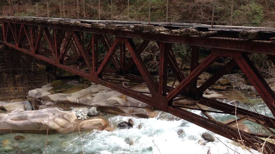 A rusted metal bridge over a fast-flowing river with rocky banks and dense forest in the background.