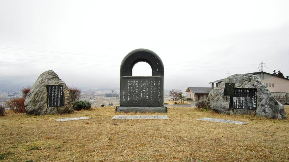 A stone monument with inscriptions, surrounded by rocks and a building in the background.