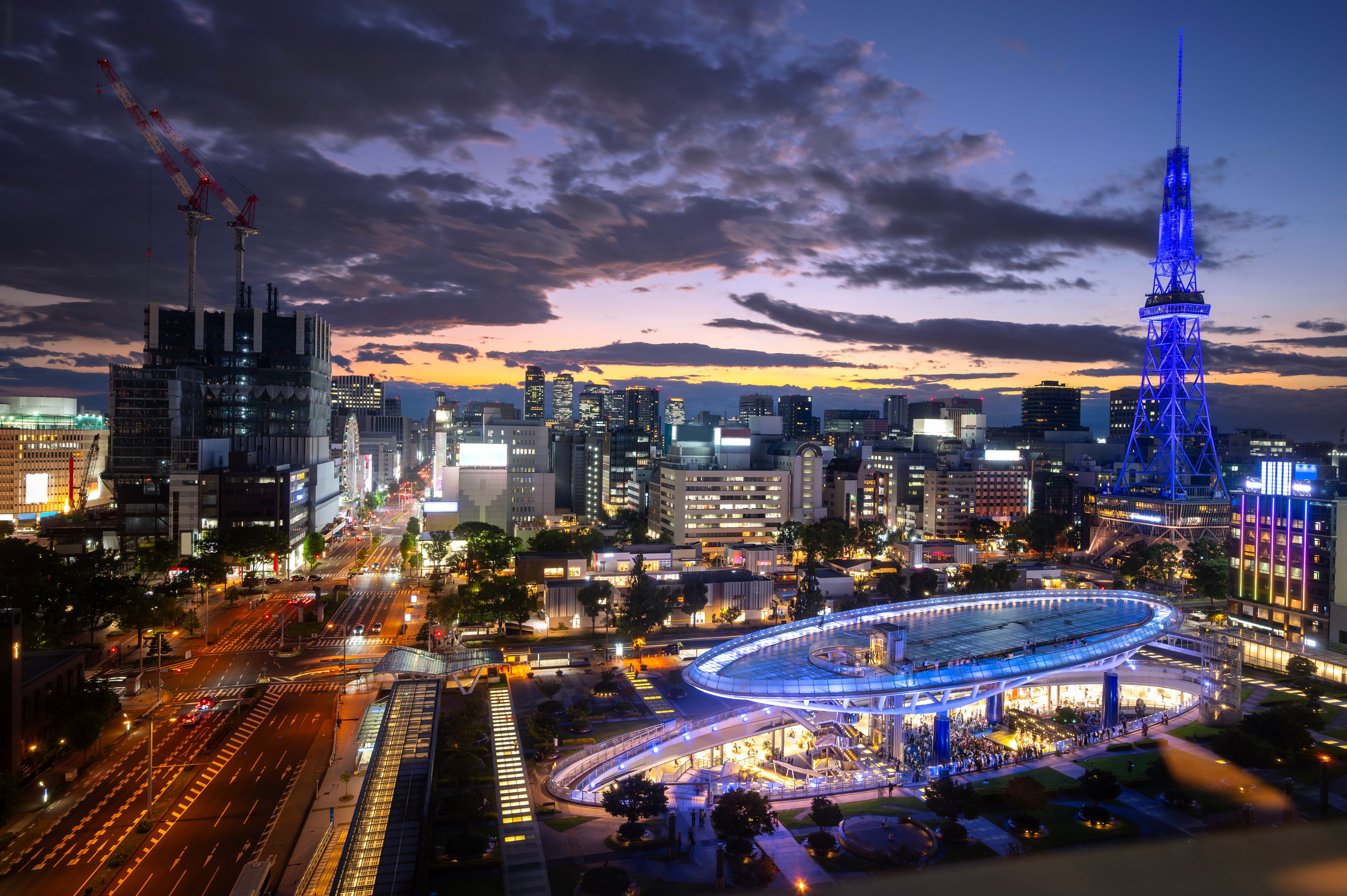 A cityscape at dusk with illuminated buildings, a prominent blue-lit tower, and a circular structure with lights.