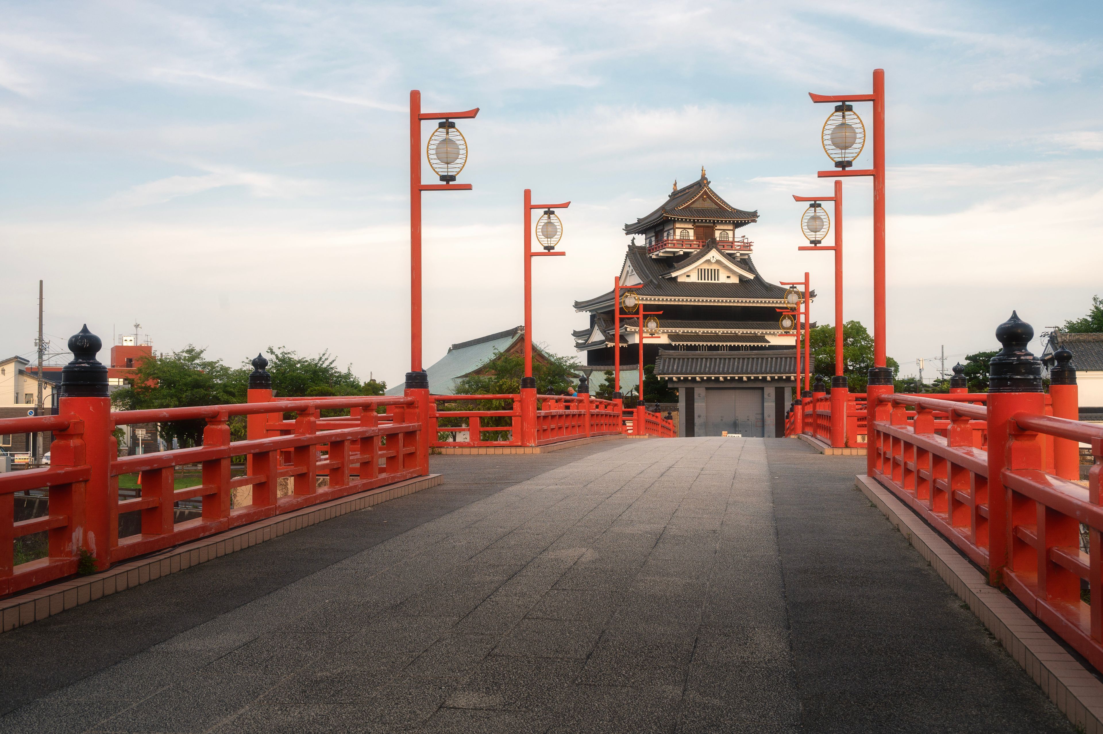 A traditional Japanese gate with red pillars and lanterns, leading to a two-tiered building with a tiled roof.