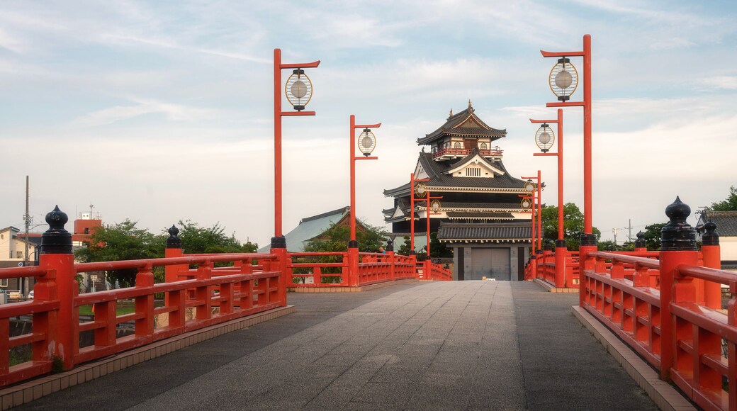 A traditional Japanese gate with red pillars and lanterns, leading to a two-tiered building with a tiled roof.