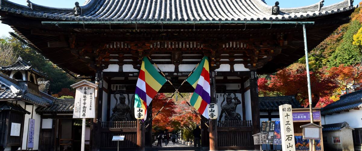 A traditional Japanese gate with two flags, surrounded by autumn foliage and traditional architecture.
