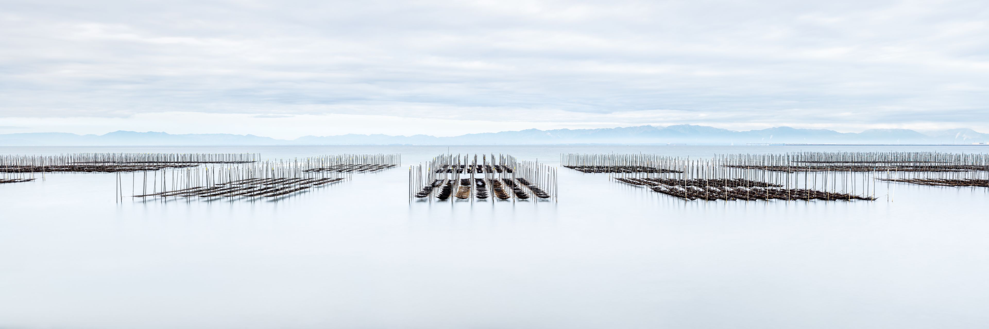 Una extensa masa de agua tranquila con una serie de estructuras de madera que se adentran en el agua, rodeada por un paisaje montañoso bajo un cielo nublado.