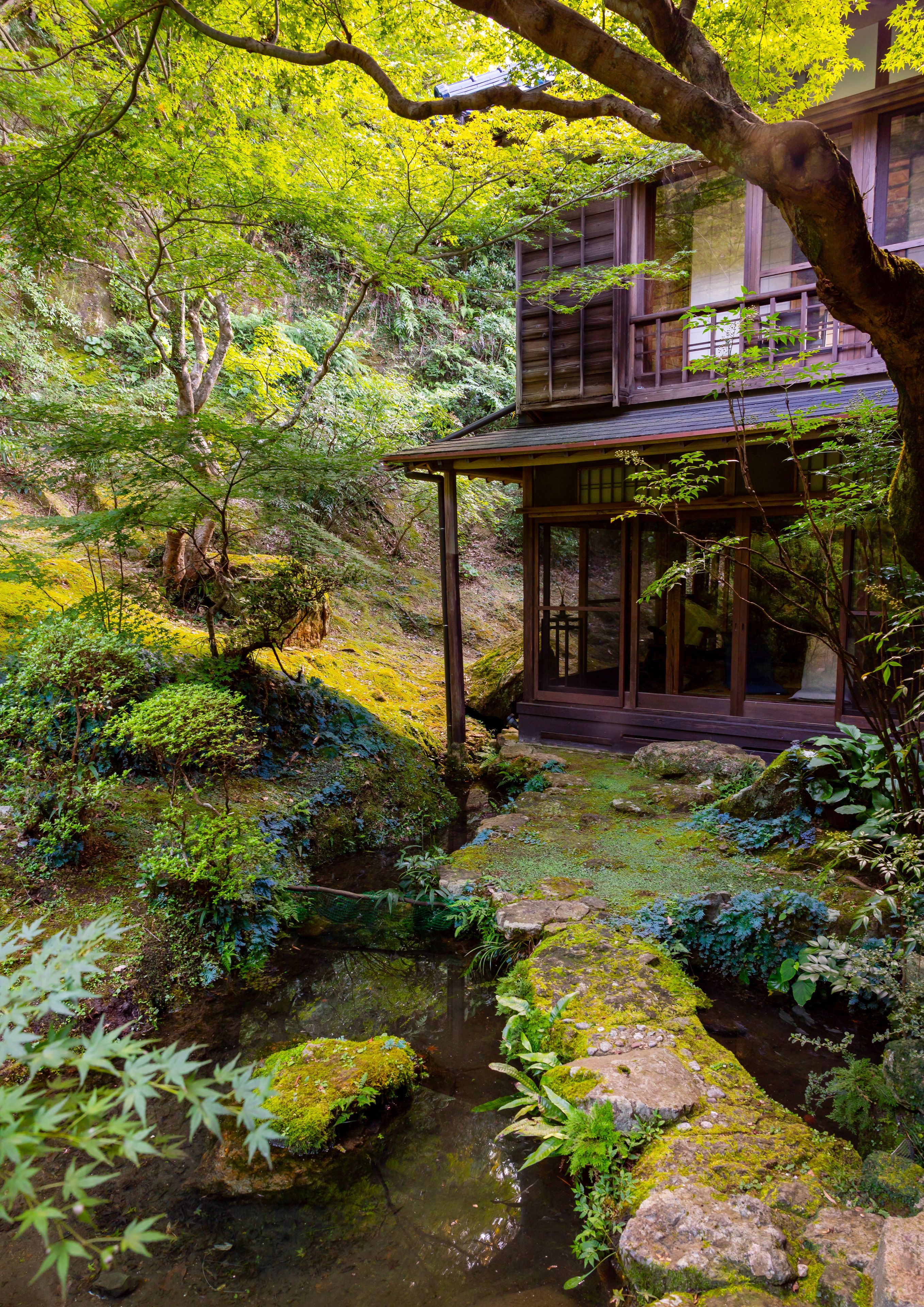 Une structure traditionnelle japonaise en bois, offrant une vue sur un jardin luxuriant, avec un petit ruisseau et diverses plantes.