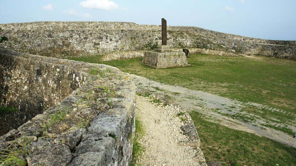 A stone fortification with a central pillar, a grassy field, and a stone wall.