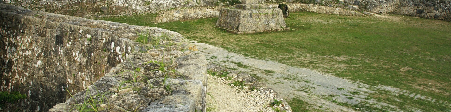 Una fortificación de piedra con un pilar central, un campo cubierto de césped y una pared de piedra.