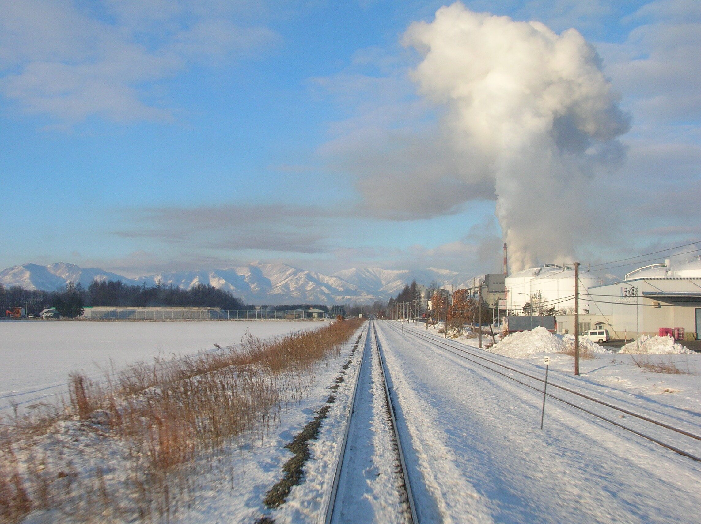 A railway track with snow-covered surroundings, a building with smoke, and snow-capped mountains in the background.