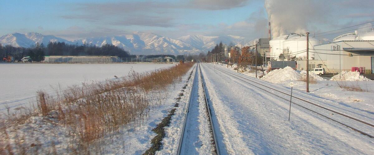 A railway track with snow-covered surroundings, a building with smoke, and snow-capped mountains in the background.