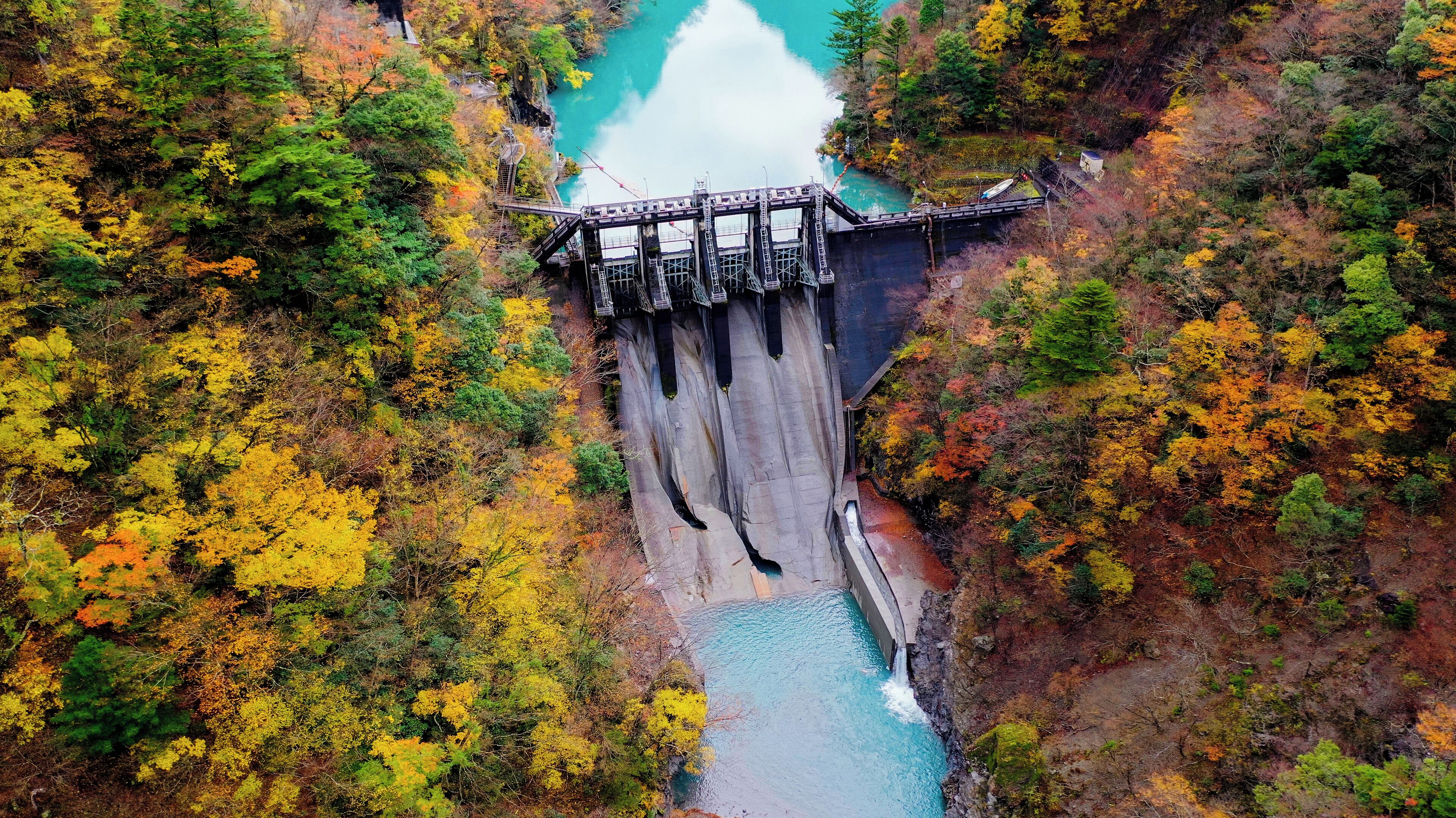 A dam with a bridge, surrounded by autumn-colored trees and a turquoise river.