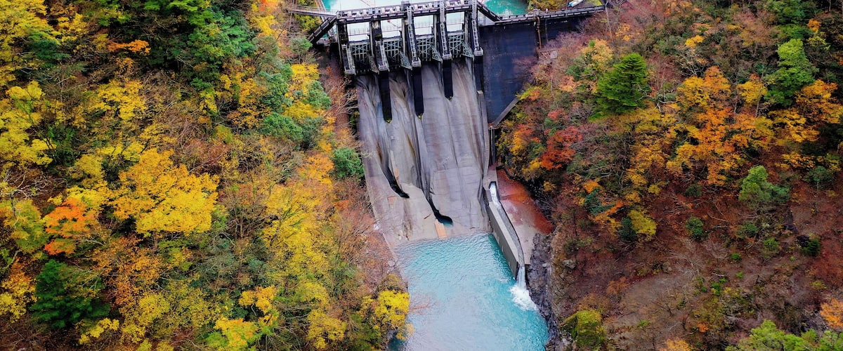 A dam with a bridge, surrounded by autumn-colored trees and a turquoise river.