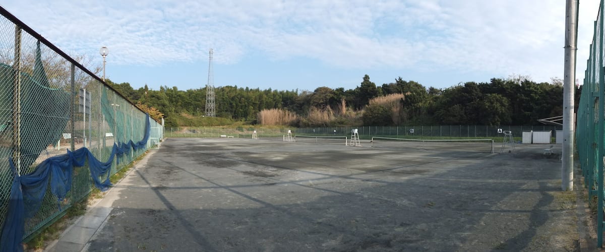 An empty sports court with a chain-link fence, a tall communication tower, and a forested hill in the background.