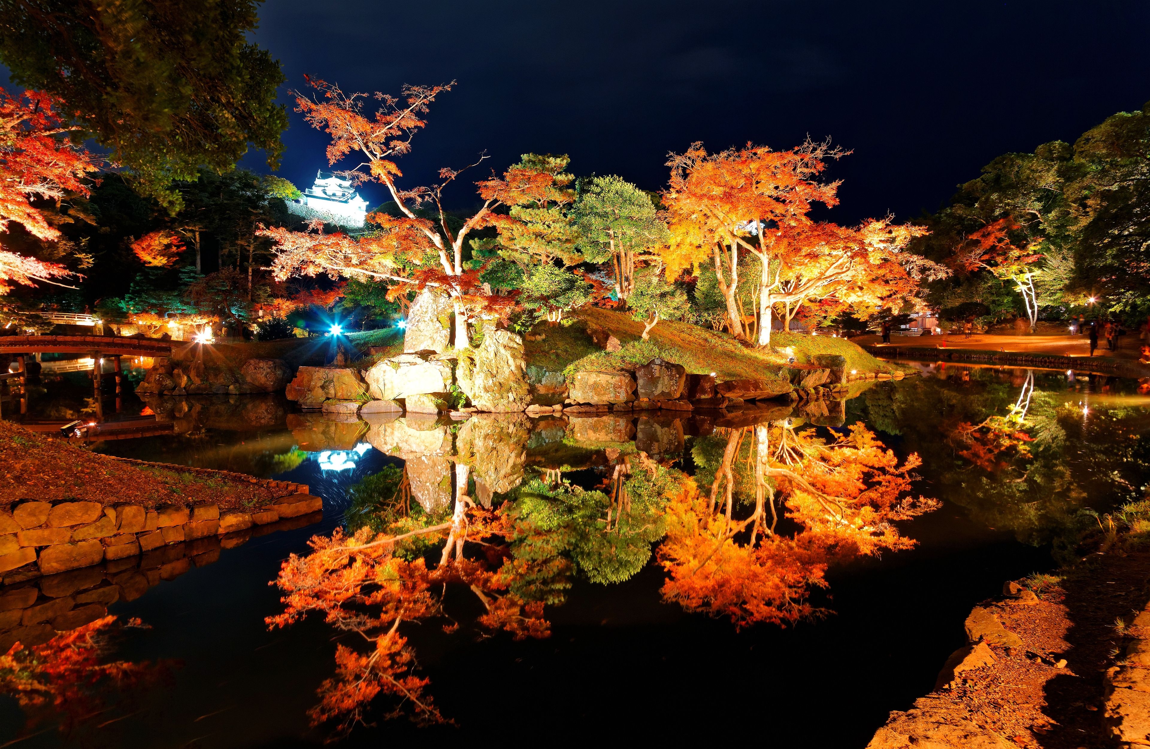 A nighttime view of a serene park with autumn-colored trees, a reflective pond, and traditional architecture.