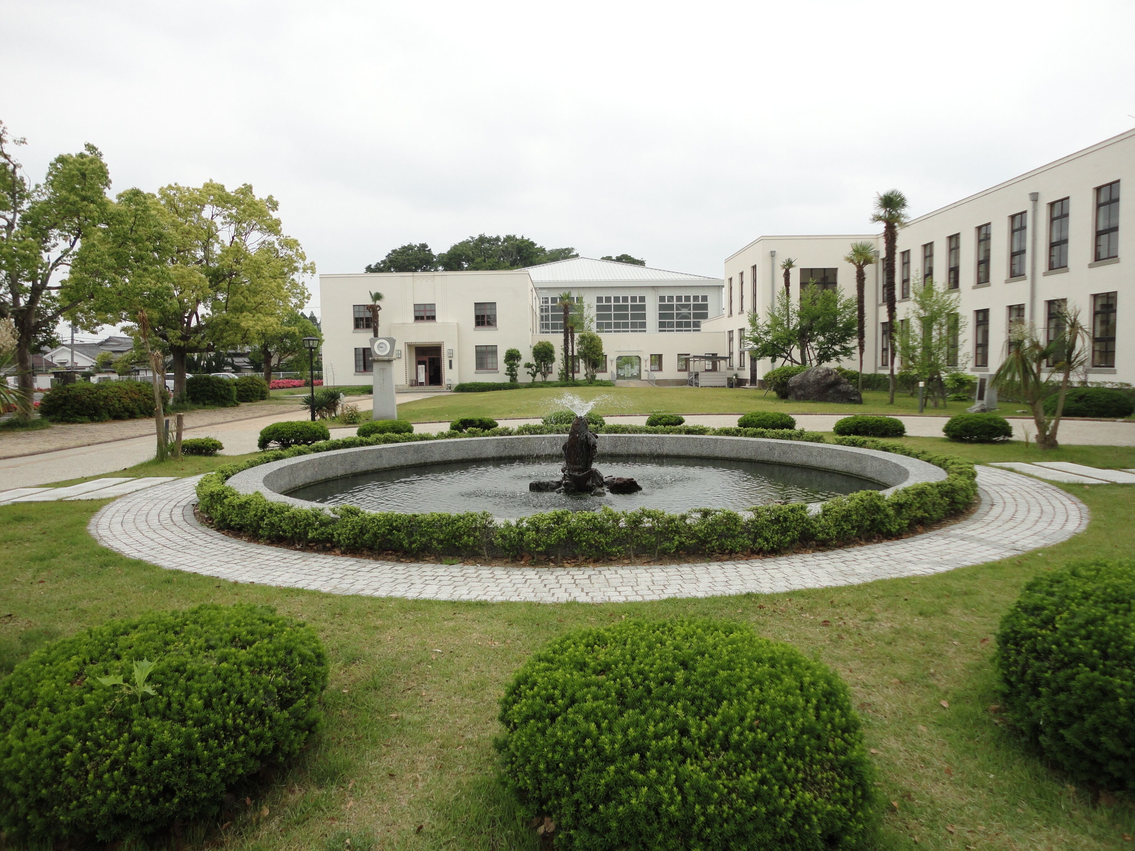 A landscaped garden with a circular fountain, trimmed bushes, and a building with multiple windows in the background.