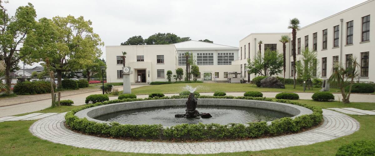 A landscaped garden with a circular fountain, trimmed bushes, and a building with multiple windows in the background.