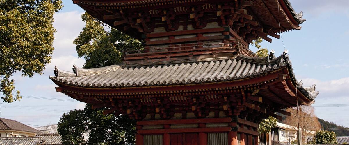 A traditional multi-tiered pagoda with intricate wooden carvings and a tiled roof, surrounded by a gravel path and a wooden fence.