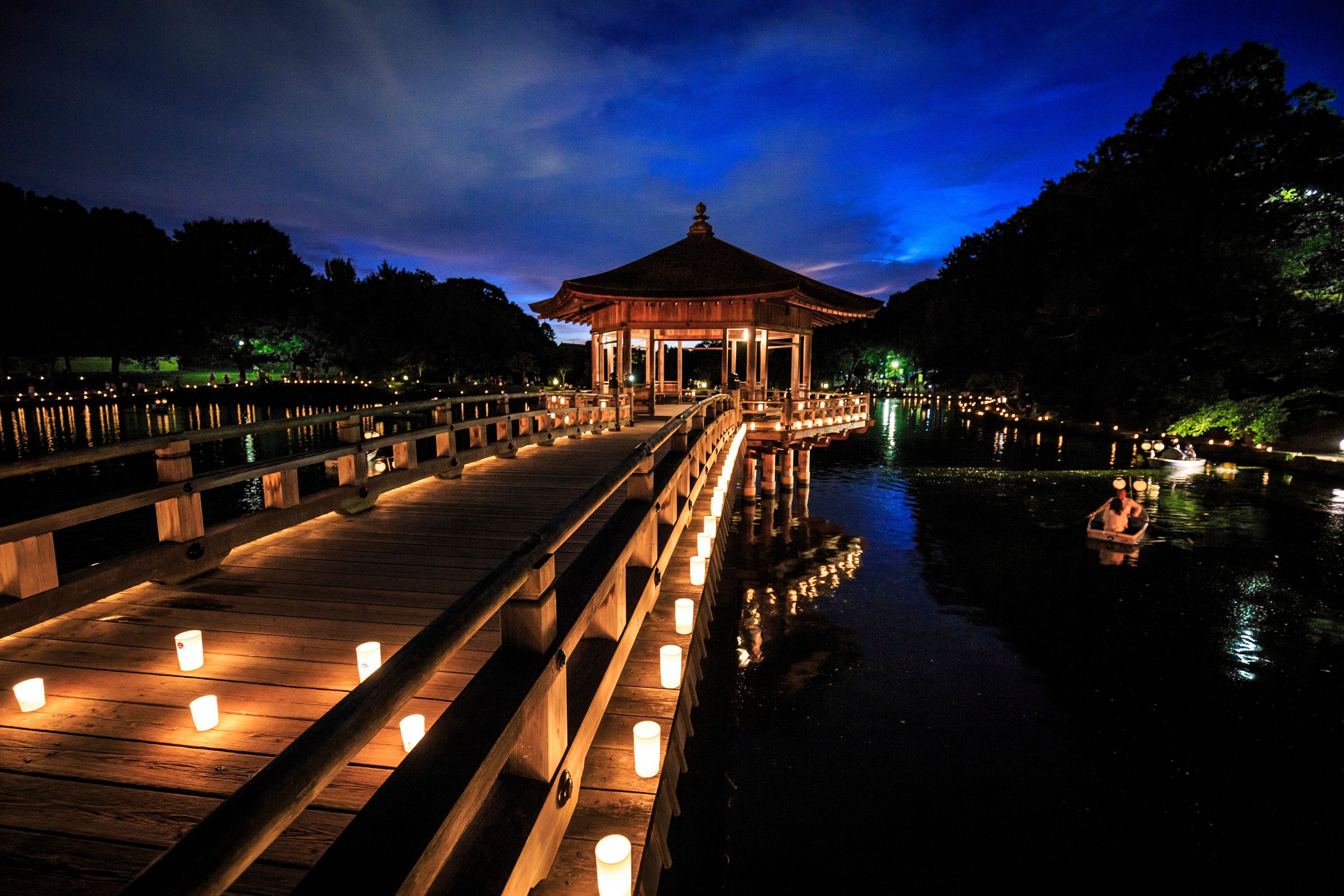 A wooden bridge with lit candles leading to a traditional pavilion by a calm body of water at night.