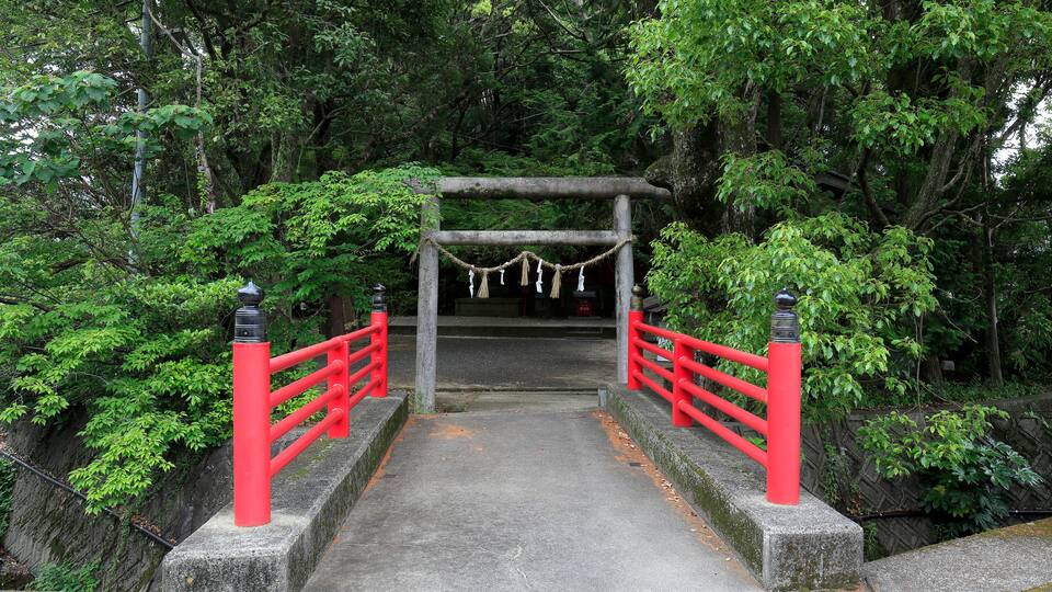 A stone bridge with red railings leading to a wooden archway surrounded by lush greenery.