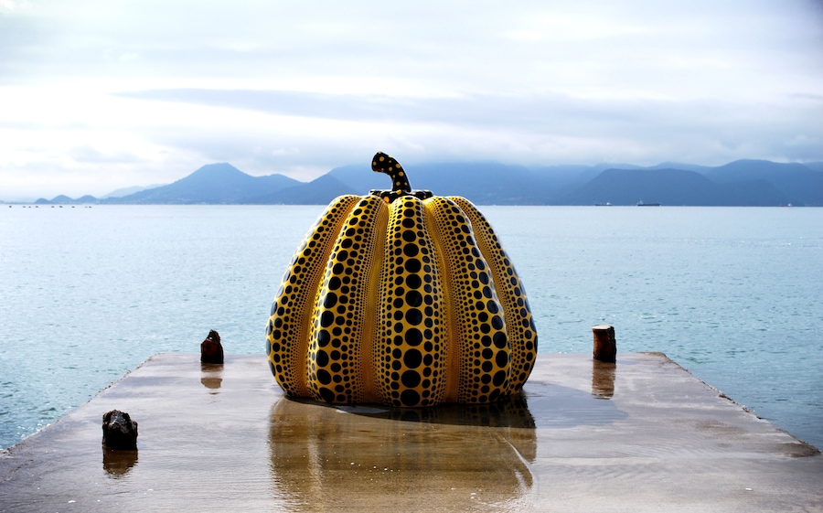 A large polka-dotted sculpture on a concrete platform by a calm body of water with mountains in the background.