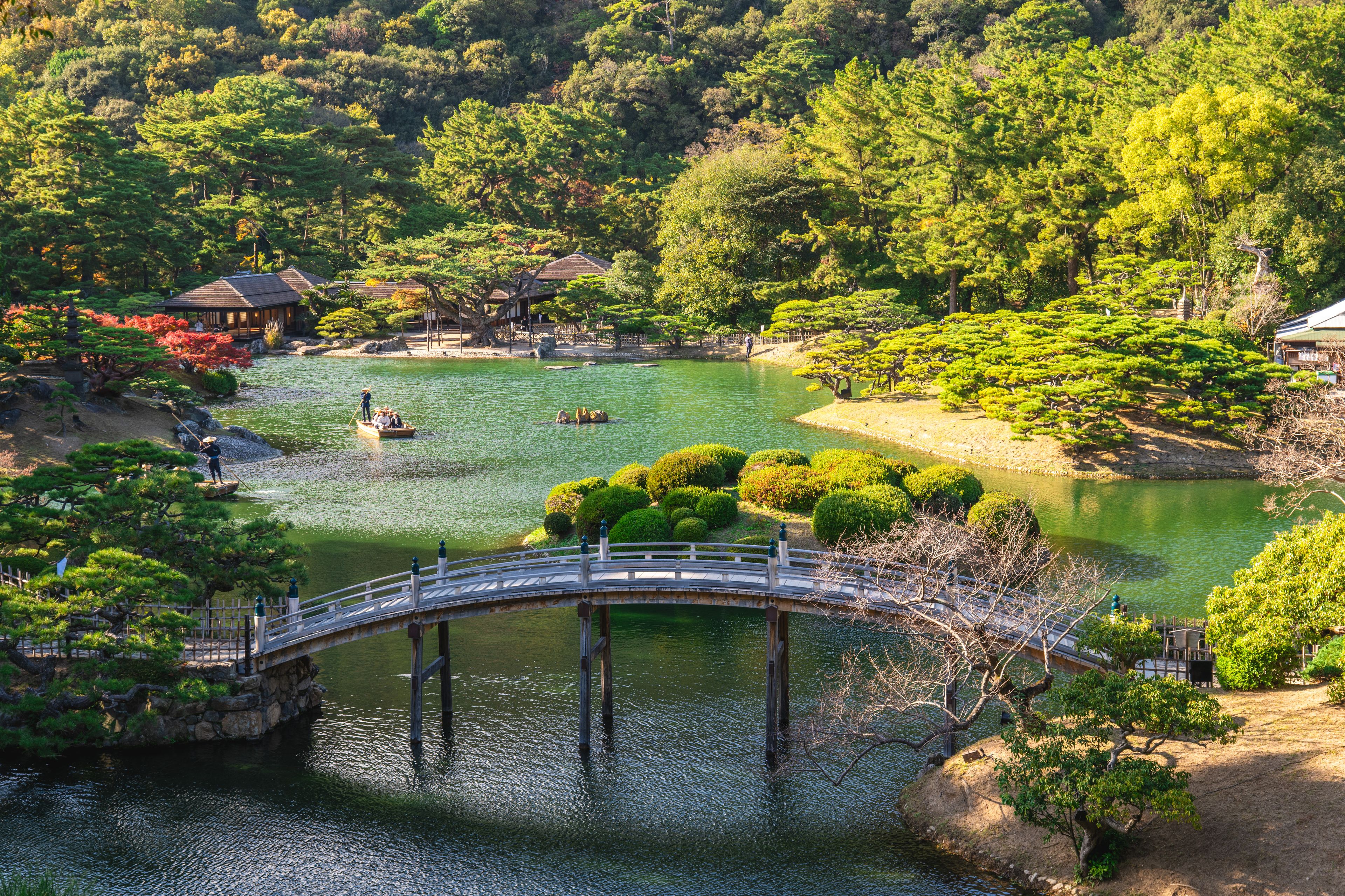 A serene Japanese garden with a bridge over a pond, surrounded by lush greenery and traditional buildings.