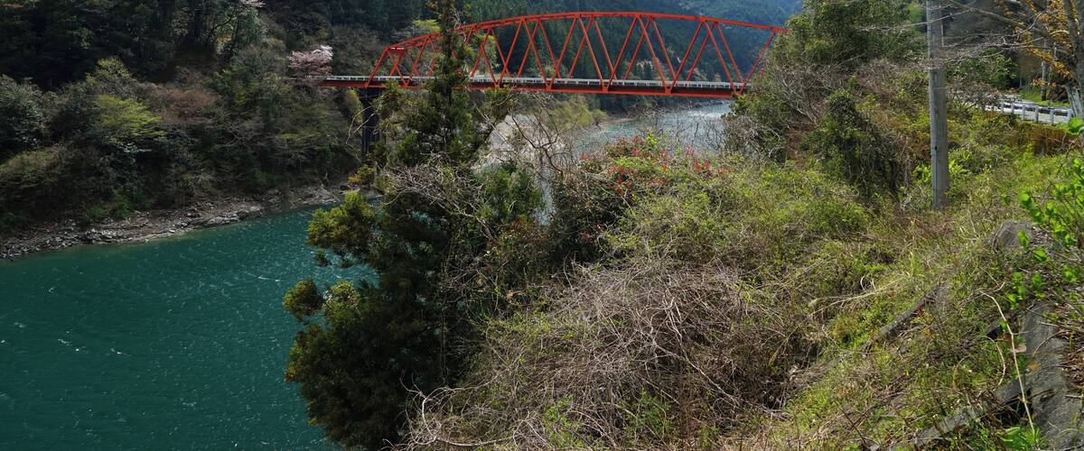 A red bridge over a river, surrounded by mountains and dense forest.
