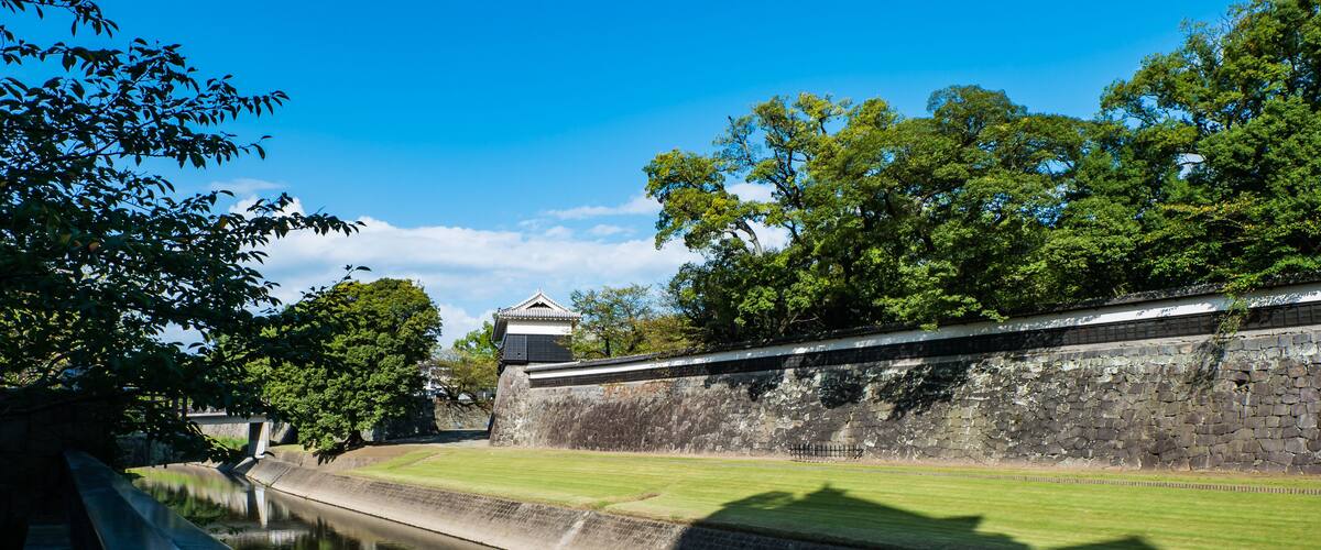 A stone wall with a traditional building, a canal, and lush green trees.
