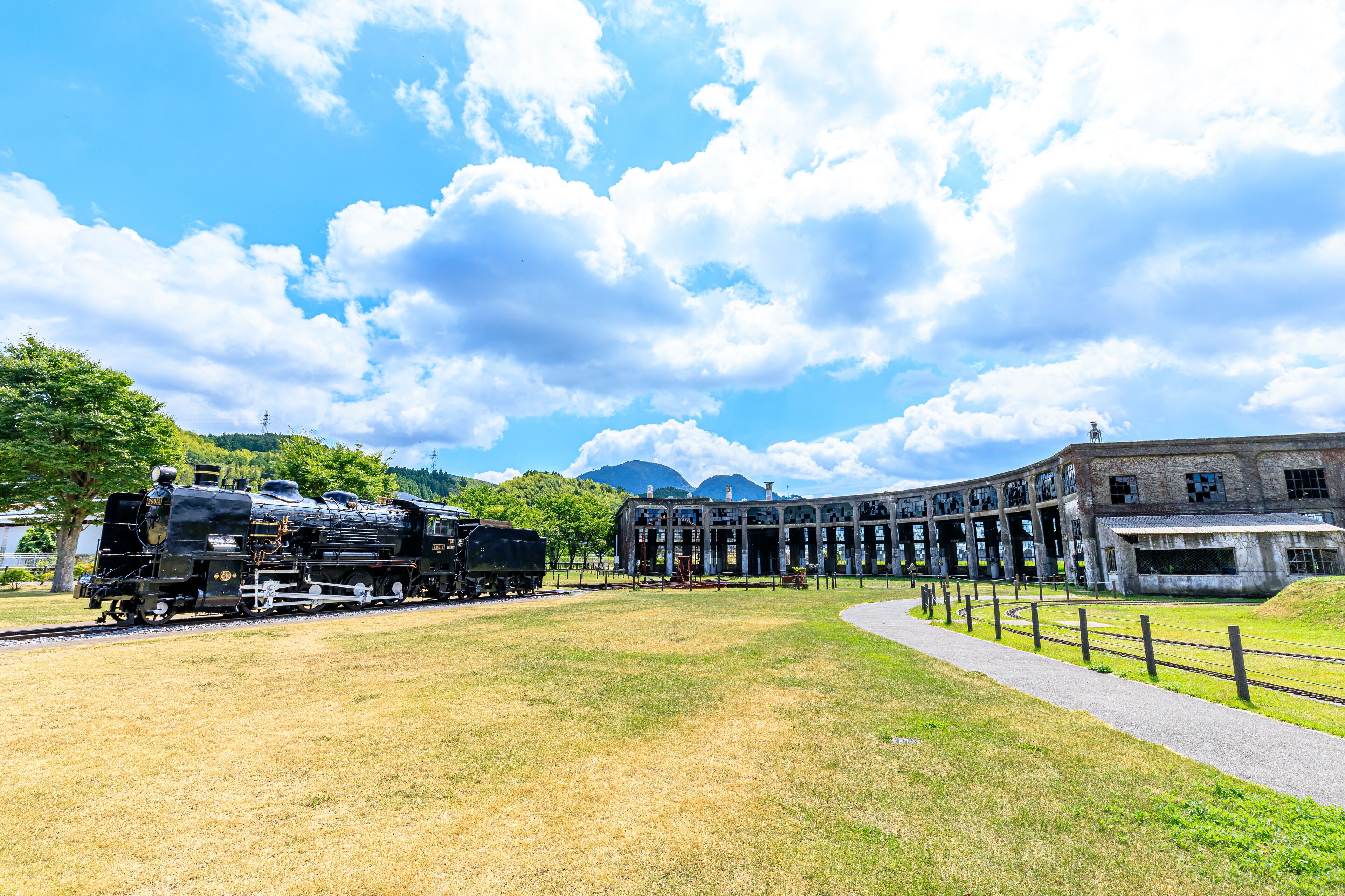 A historic train station with a steam locomotive, a large building with a curved roof, and a grassy field.