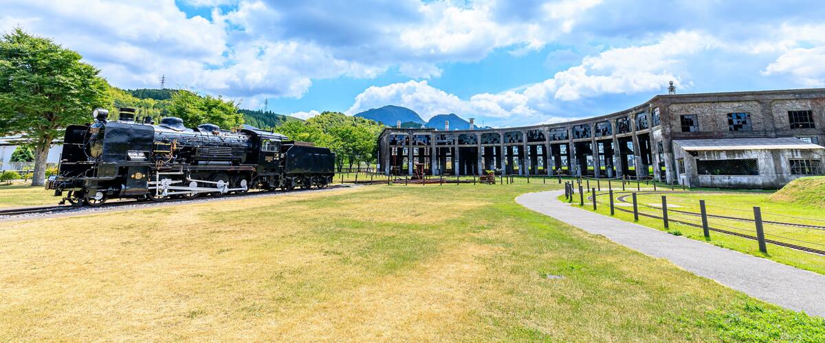 A historic train station with a steam locomotive, a large building with a curved roof, and a grassy field.