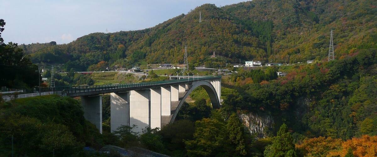 A large concrete bridge spanning a river, surrounded by dense forest and hills.