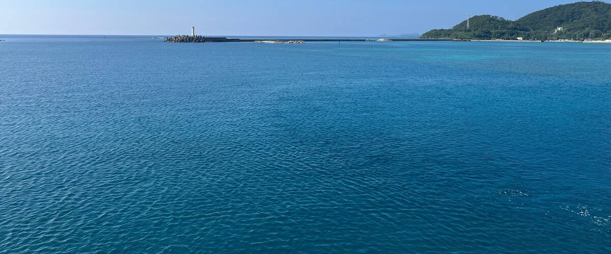 A calm sea with a lighthouse and a green hill in the distance.