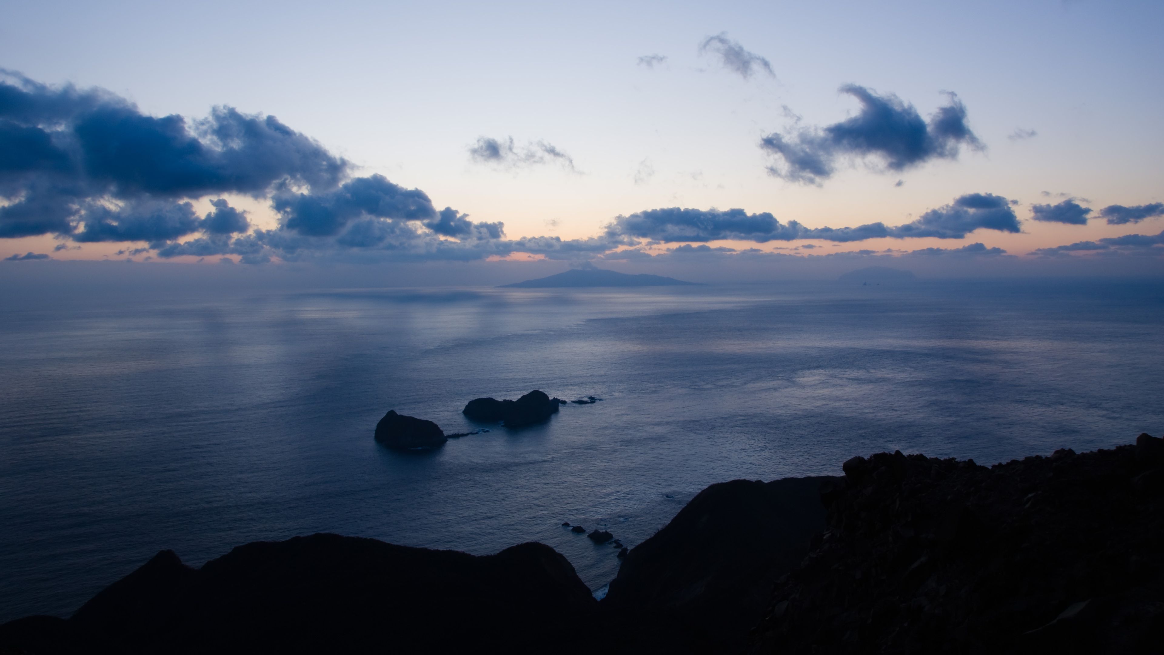 A coastal landscape with calm waters, rocky outcrops, and a distant mountain range under a twilight sky.