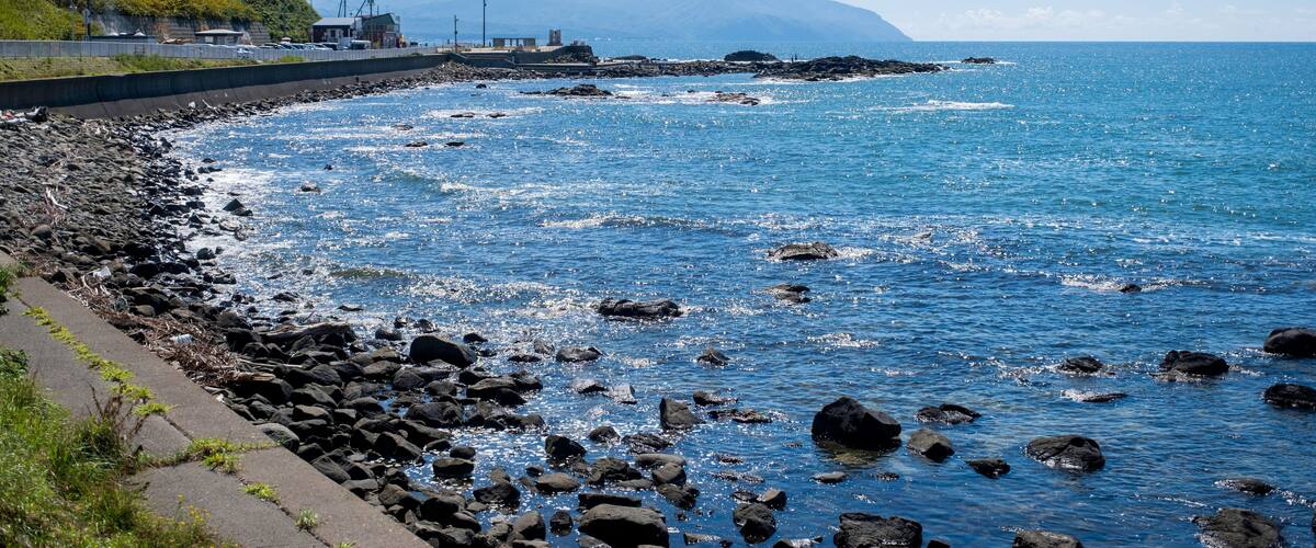 A rocky shoreline with a clear blue sea, a distant mountain, and buildings on a hill.