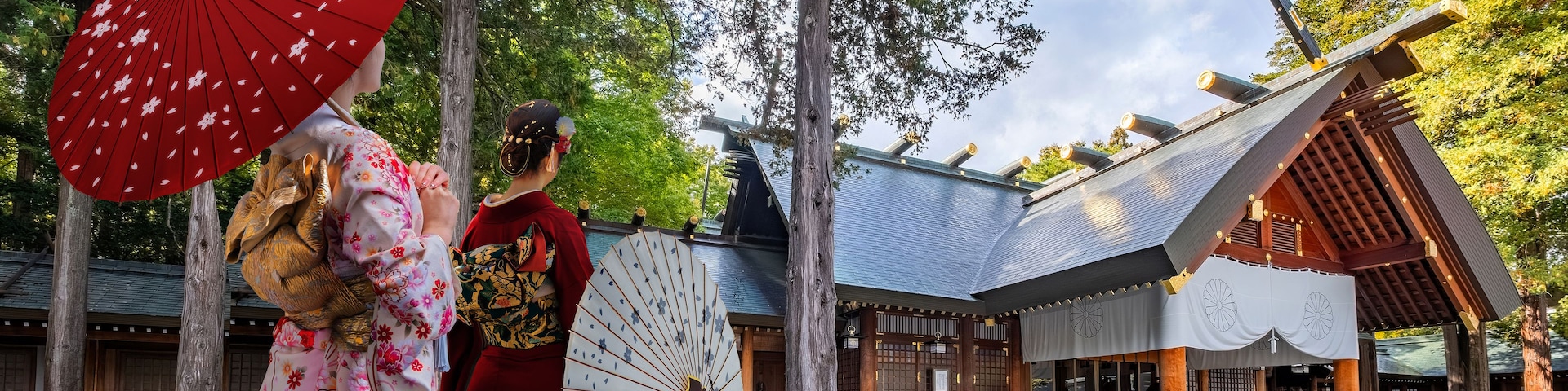 Edifício tradicional japonês com telhado inclinado e vigas de madeira, cercado por árvores e um caminho de cascalho.